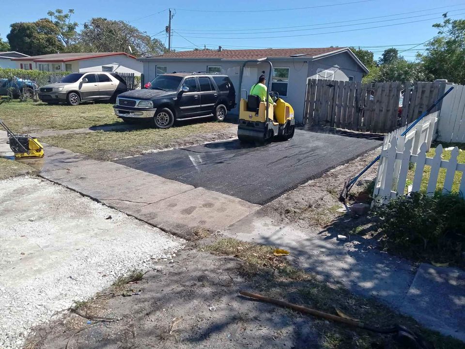 A person operates a small yellow steamroller to perform asphalt driveway paving in front of a house, with parked cars and a white picket fence nearby on a sunny day in Tampa FL.