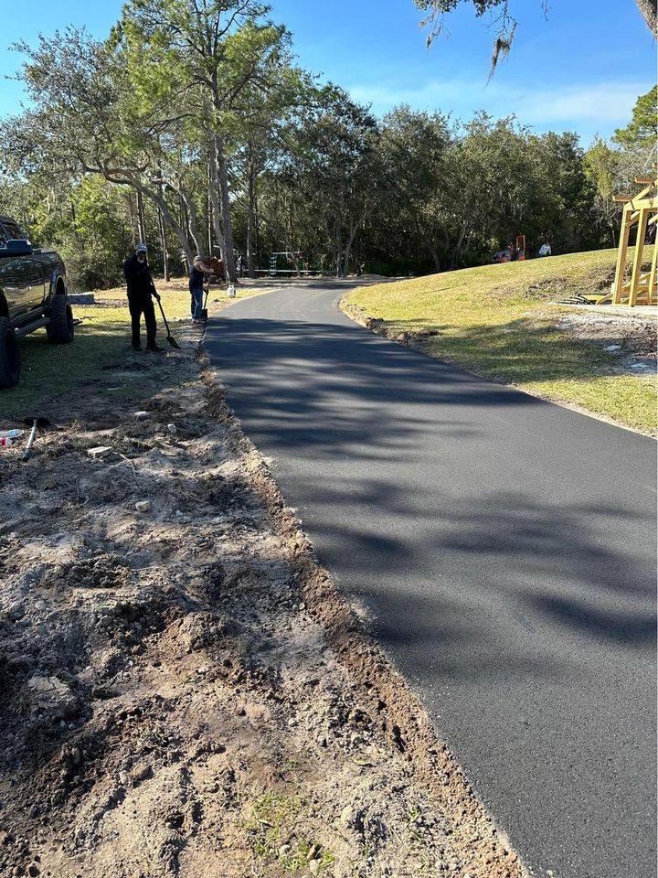 Workers stand near a freshly paved asphalt path, showcasing expert paving services in a grassy, tree-lined area under Tampa FL's clear blue sky. A truck is parked on the left, with construction materials for asphalt driveway paving on the right.