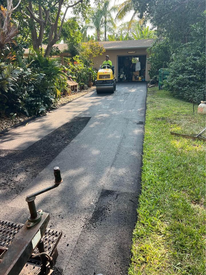 A worker operates a small steamroller to smooth fresh asphalt on a driveway leading to a garage, surrounded by green plants and trees—a sunny day perfect for Asphalt Driveway Paving and expert paving services in Tampa FL.