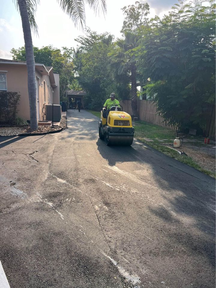 A person drives a yellow asphalt roller on a freshly paved driveway between houses, showcasing expert asphalt paving services in Tampa FL, surrounded by trees and greenery on a sunny day.
