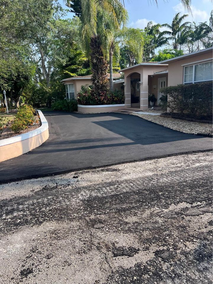 A house in Tampa FL with beige walls and a new, smooth black asphalt driveway. The yard features palm trees, bushes, and stone landscaping. The street in front shows a rough surface—perfect for asphalt paving services or resurfacing.