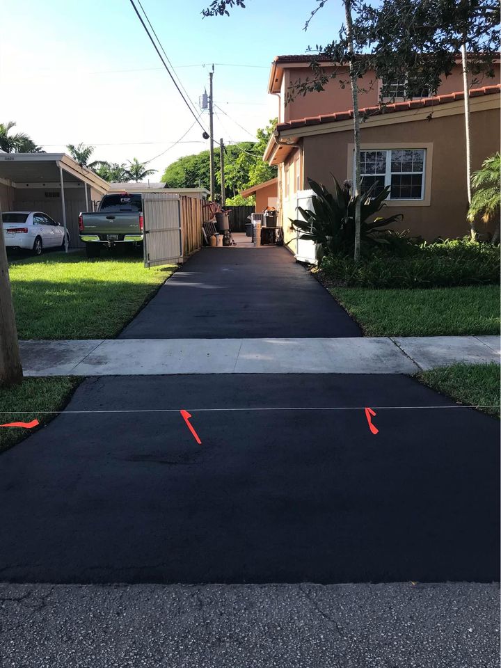 A freshly paved asphalt driveway leads to a tan house in Tampa, FL, with red arrows marking where the new surface meets the sidewalk. A truck is parked near a fenced area beside the house, and green grass borders the driveway.
