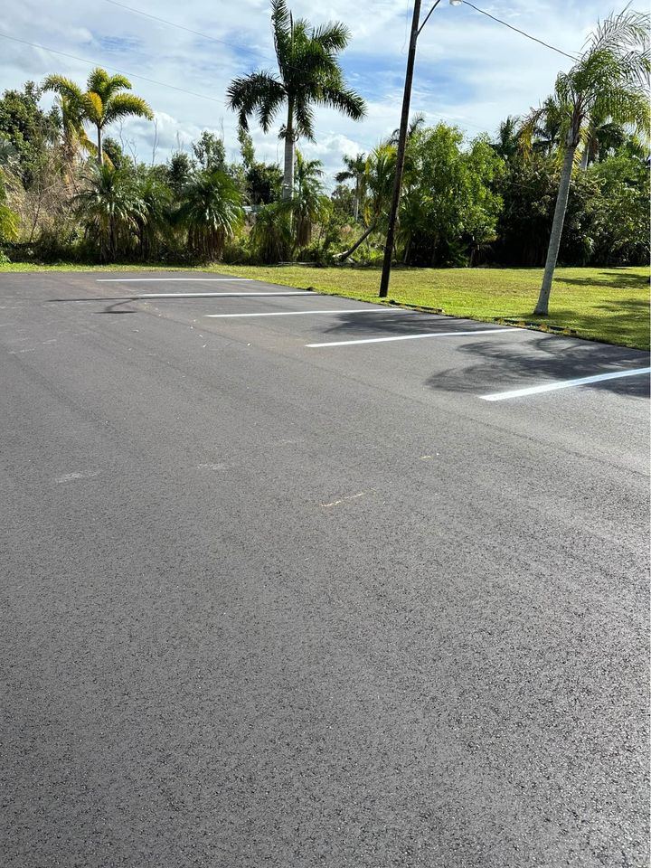 Freshly paved empty parking lot with crisp white lines, expertly maintained with recent sealcoating. Bordered by palm trees and green grass under a partly cloudy sky.