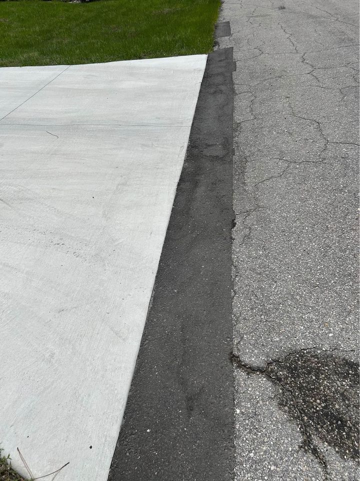 A concrete driveway meets an asphalt strip, which borders a cracked asphalt road in need of asphalt repair Tampa. Green grass accents the upper left corner near the driveway.