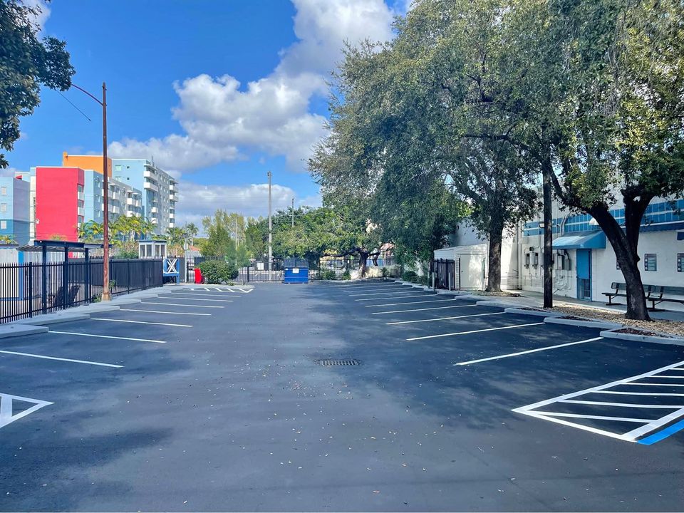 A freshly paved asphalt parking lot with crisp white and blue-lined parking lot striping, surrounded by trees and buildings under a blue sky with scattered clouds in Tampa FL.