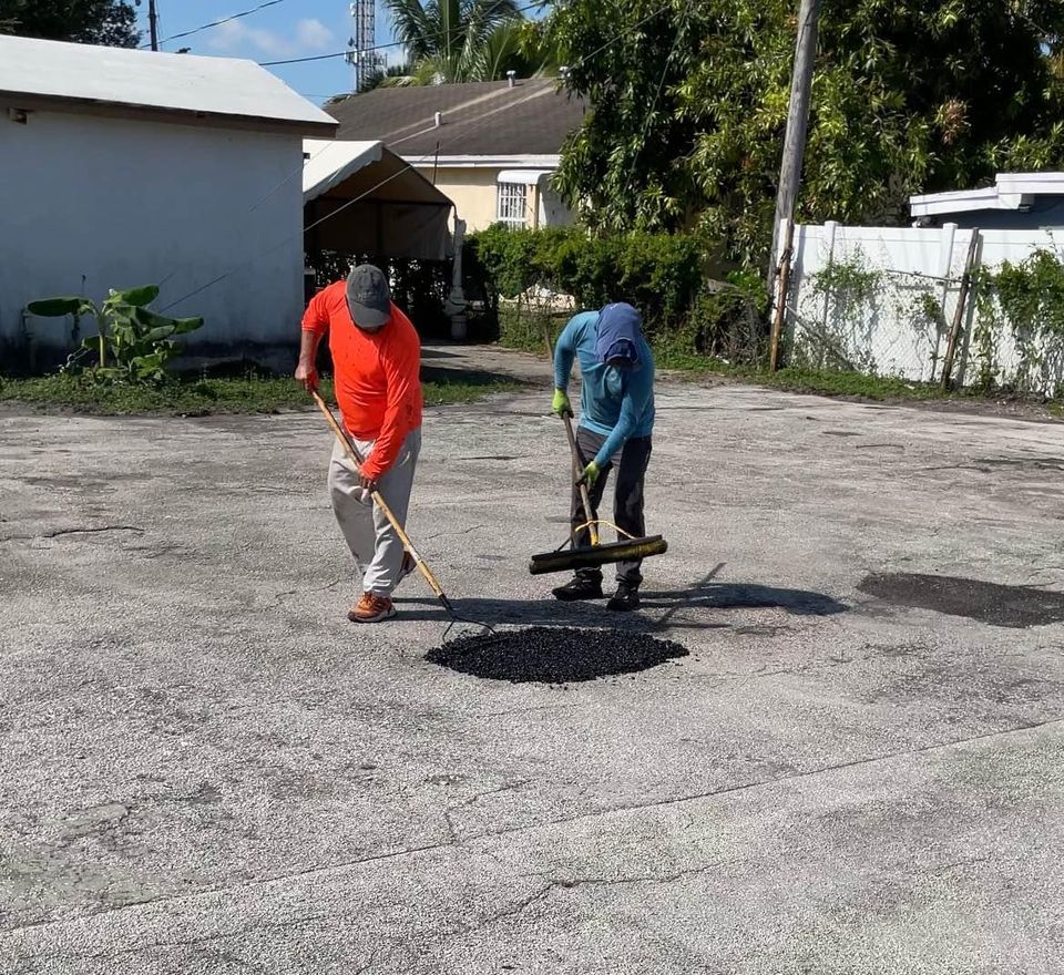 Two workers fill and smooth a pothole in an asphalt surface using shovels and tools on a sunny day. This scene highlights expert asphalt driveway repair, with trees and buildings visible in the background.