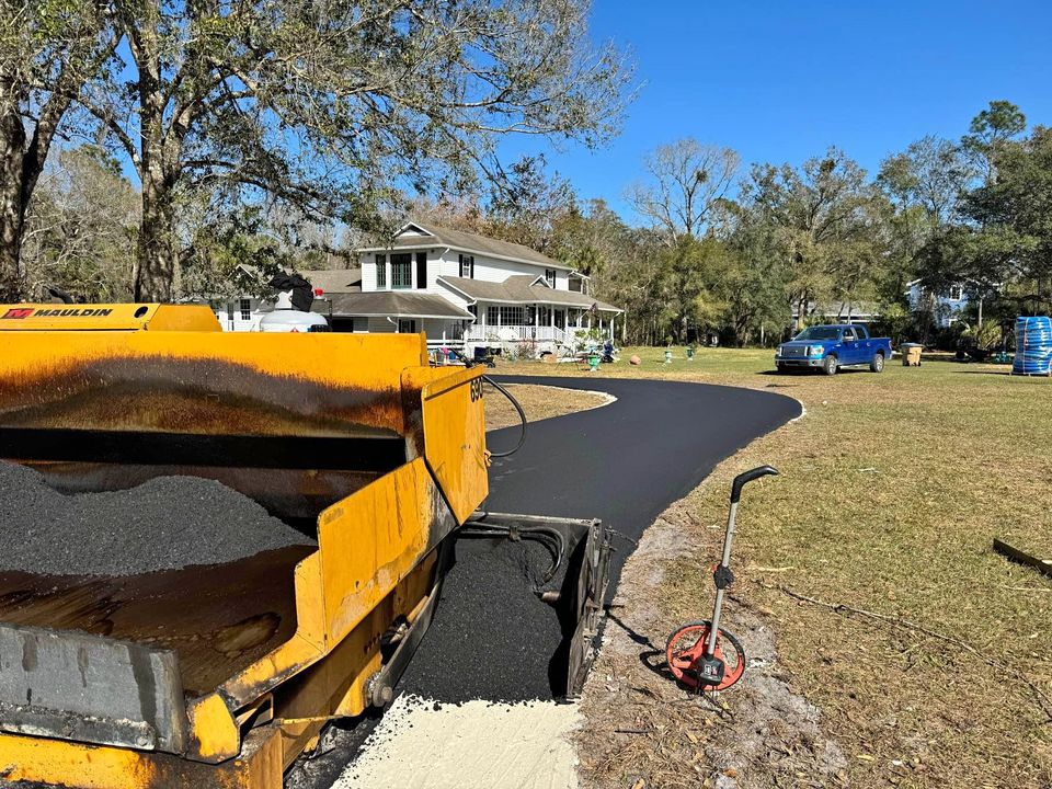 A yellow asphalt paving machine lays a fresh black driveway leading to a two-story house in Tampa FL, surrounded by trees and lawn under a clear blue sky. A blue vehicle and paving services materials are visible in the yard.