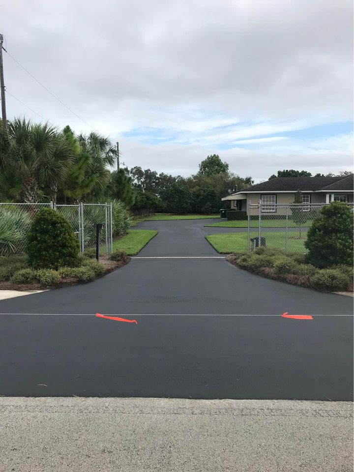 A paved asphalt driveway leads to a house surrounded by green lawns and bushes, with a chain-link fence and gate on either side. Two orange arrows are marked at the entrance. For sealcoating services in Tampa FL, this home makes a great impression under cloudy skies.