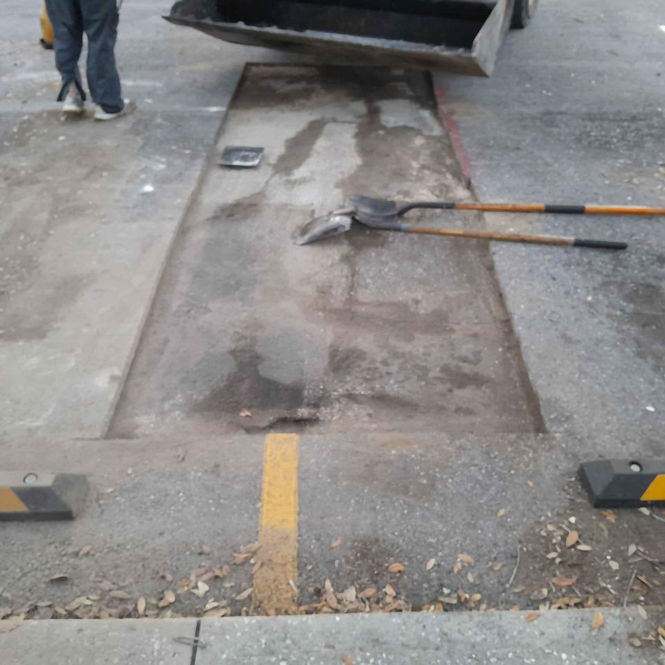 A shovel and a rake lie on a patch of road with a yellow line misaligned across a repaired section, hinting at recent paving work. A worker stands nearby, providing asphalt parking lot maintenance, while a truck bed is visible in the background.
