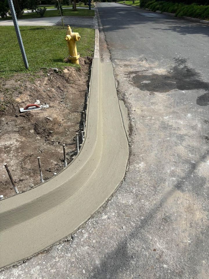 Freshly poured concrete forms a new sidewalk ramp curving from the street onto the grass. A yellow fire hydrant stands nearby, with construction tools and rebar visible—an example of paving services alongside asphalt parking lot maintenance.