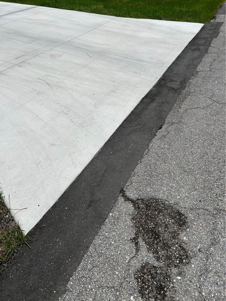 A section of clean, light-colored concrete driveway meets older, cracked asphalt pavement in need of asphalt parking lot maintenance, with a dark oil stain visible on the asphalt. Green grass borders the edge of the driveway.