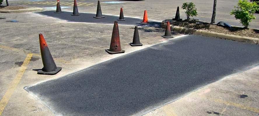 Several orange traffic cones surround a freshly paved rectangular patch of asphalt in a parking lot, marking off the area from vehicles—a clear sign of recent asphalt parking lot maintenance. Shrubs and trees are nearby.