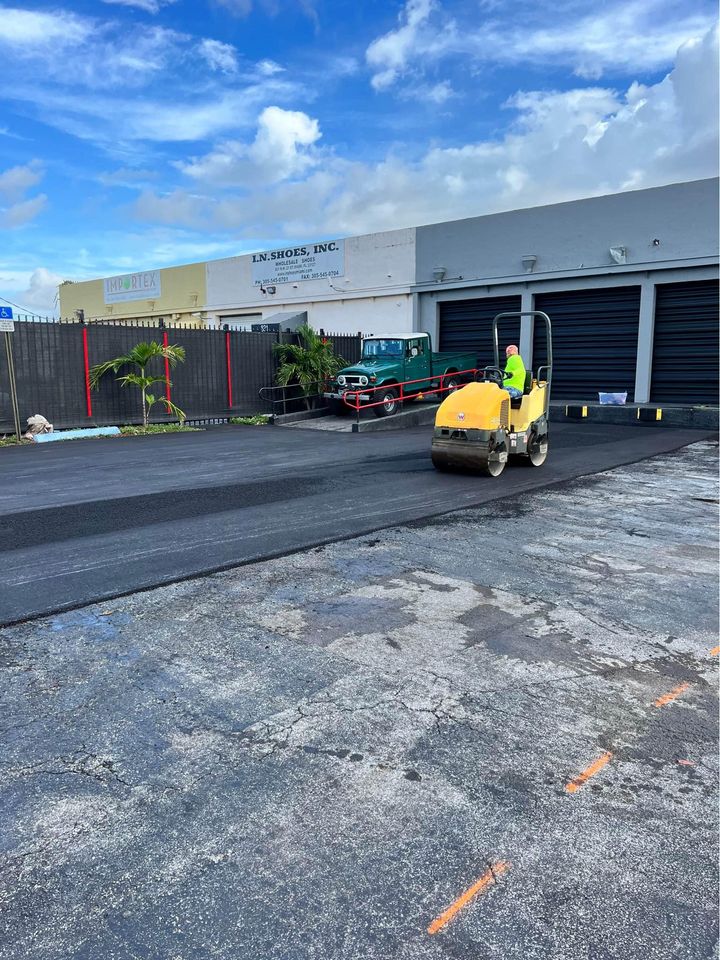 A worker in a yellow vest operates a steamroller, providing professional paving services to ensure quality asphalt parking lot maintenance beside an industrial building under a partly cloudy blue sky.