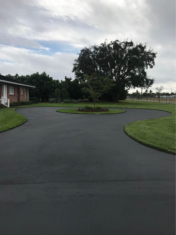 A freshly paved asphalt driveway with a green grassy island and a small tree in the center sits in front of a house and large trees under a cloudy sky—showcasing quality sealcoating services in Tampa FL.