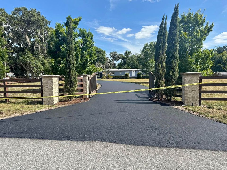 A paved asphalt driveway leads to a house behind a wooden fence and stone pillars. Yellow caution tape blocks the open entrance, possibly for sealcoating services. Surrounded by trees and under a blue Tampa FL sky, the area remains inaccessible.