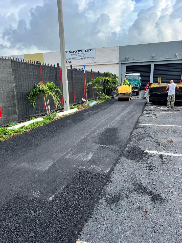 Workers are paving a driveway with fresh asphalt next to a building in Tampa FL, using a green and yellow road roller. The fenced area features small palm trees along the side, with cloudy skies above—perfect for asphalt paving of parking lots.