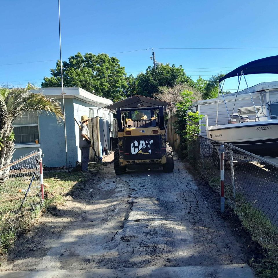 A construction worker stands beside a yellow CAT backhoe parked on a cracked asphalt driveway ready for resurfacing, between a house and a boat on a trailer. On this sunny Tampa day, the fenced area is surrounded by lush greenery.