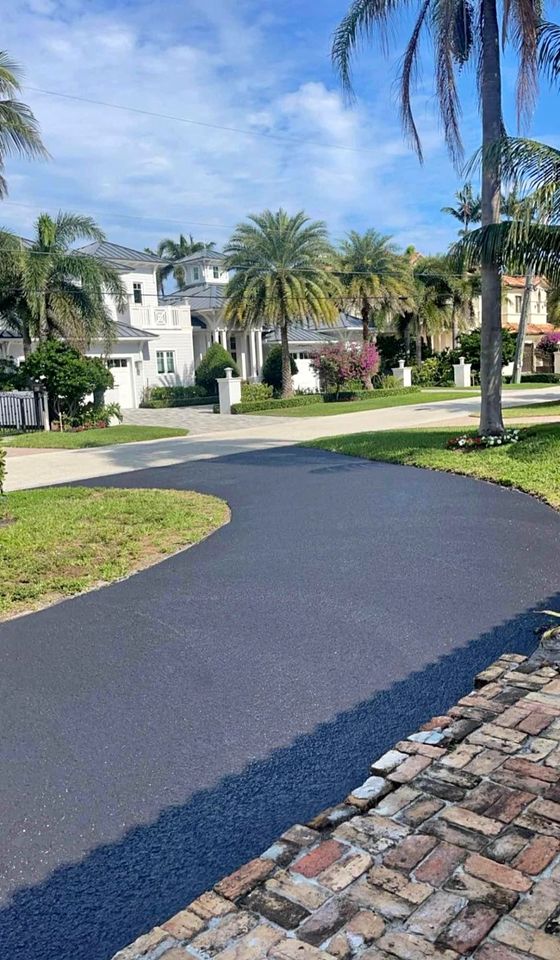 A newly paved black asphalt driveway curves through a green lawn towards white houses surrounded by palm trees on a bright, sunny day, showcasing quality paving services Tampa Bay residents trust.